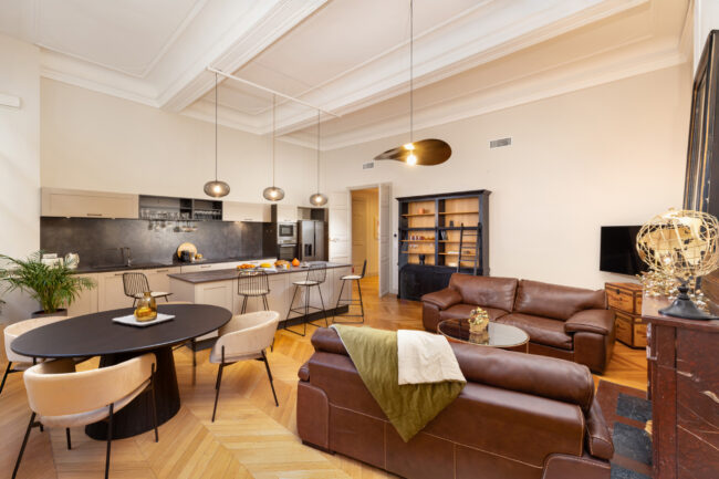 Vaste salon en plan large avec deux canapés en cuir marron, une table noire des chaises beige et un ilot cuisine en arrière plan,