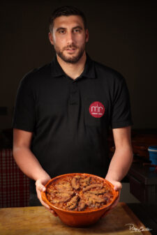 Un homme en tee shirt noir tient dans ses mains une terrine de Cassoulet gratiné.
