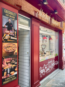 Façade rouge de trois quart de la boucherie rabat avec en premier plan une pancarte verticale faisant la promotion du cassoulet