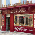 Façade rouge de la boucherie Michel Rabat avec une pancarte cassoulet fait maison et une pancarte conserves artisanales