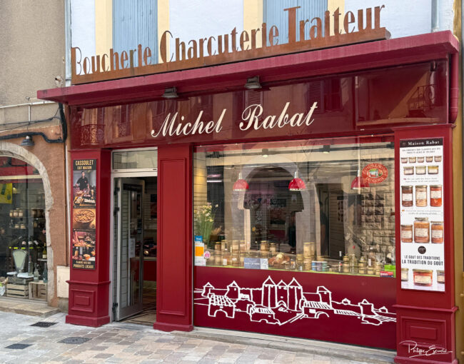 Maison Rabat, Photos + affiches + panneaux = communication gagnante 1 Façade rouge de la boucherie Michel Rabat avec une pancarte cassoulet fait maison et une pancarte conserves artisanales