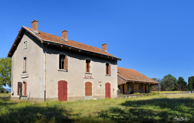 Vue large d’une gare fantôme en ruine avec prairie au premier plan.