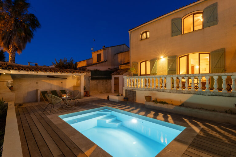 piscine éclairée de nuit avec une maison derrière et deux chaise et une table sur la gauche
