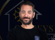 visage d'un homme barbu souriant avec un logo derrière lui en arrière plan sombre