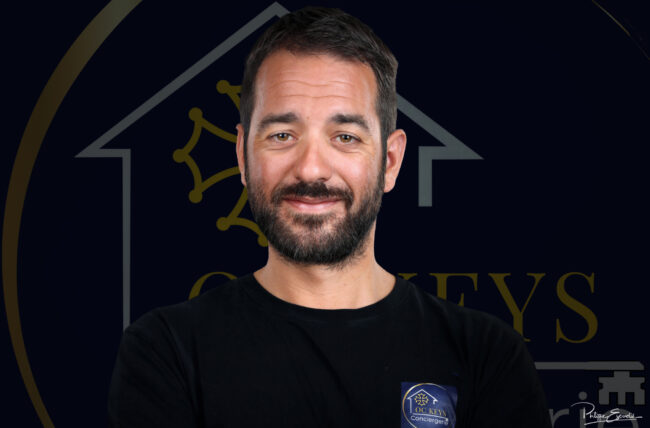 visage d'un homme barbu souriant avec un logo derrière lui en arrière plan sombre