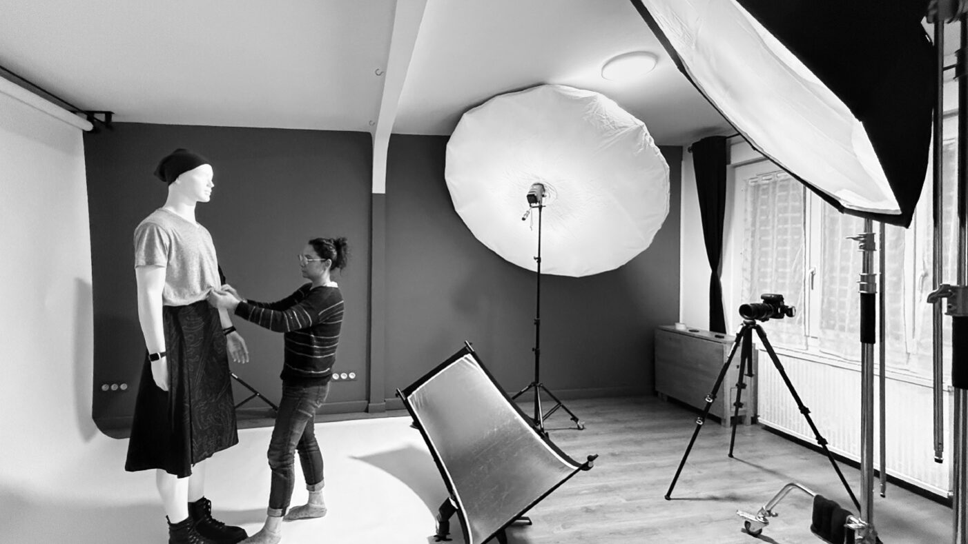 Photo noir et blanc de La créatrice Vanessa Batard qui ajuste une jupe homme sur un mannequin dans un studio photo avec des projecteurs.