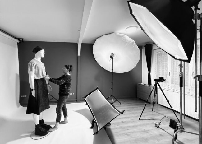 Photo noir et blanc de La créatrice Vanessa Batard qui ajuste une jupe homme sur un mannequin dans un studio photo avec des projecteurs.