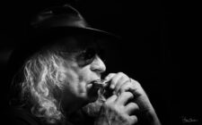 Portrait en noir et blanc d’un musicien aux cheveux longs et chapeau noir jouant de l’harmonica, photographié en gros plan avec une lumière contrastée.