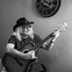 Portrait d’un musicien souriant avec chapeau et lunettes de soleil, jouant de la guitare acoustique devant un mur décoré d’une grande horloge.