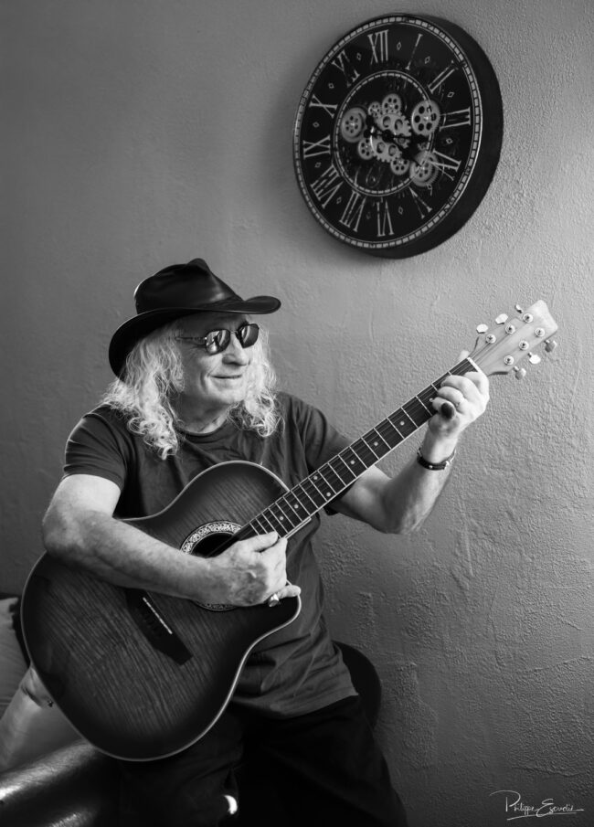 Portrait d’un musicien souriant avec chapeau et lunettes de soleil, jouant de la guitare acoustique devant un mur décoré d’une grande horloge.