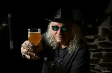 Portrait d’un homme aux cheveux longs et chapeau noir, portant des lunettes teintées, levant un verre de bière ambrée vers l’objectif dans une ambiance conviviale.