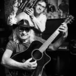Scène en studio en noir et blanc, un musicien avec guitare souriant et un photographe tenant un clap de cinéma, ambiance complice et créative.