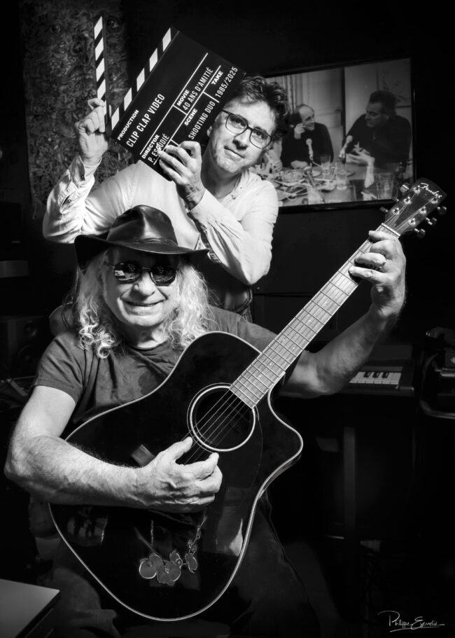Scène en studio en noir et blanc, un musicien avec guitare souriant et un photographe tenant un clap de cinéma, ambiance complice et créative.
