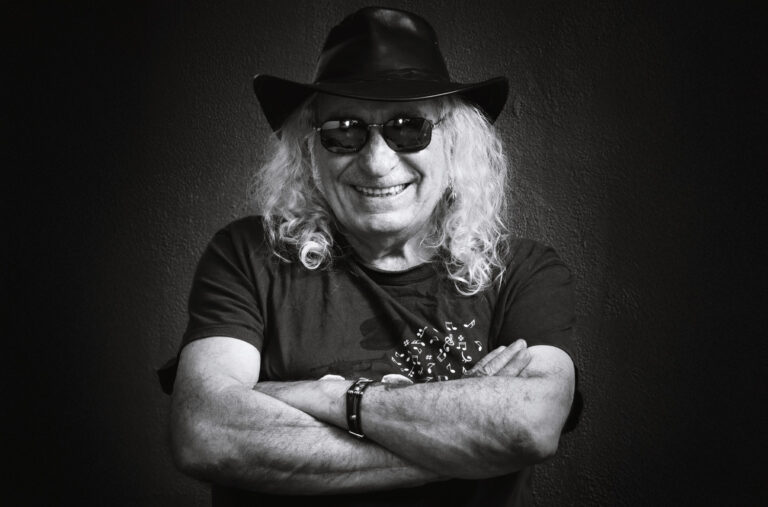 Portrait d'un homme bras croisés souriant avec un tee shirt, des lunettes de soleil et un chapeau sur fond sombre