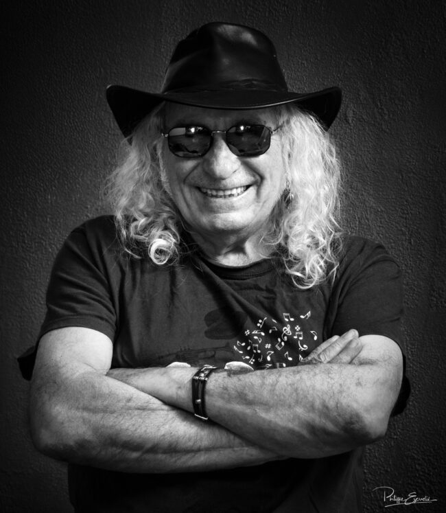 Portrait en noir et blanc d’un musicien aux cheveux longs, portant chapeau et lunettes de soleil, posant bras croisés avec un large sourire.