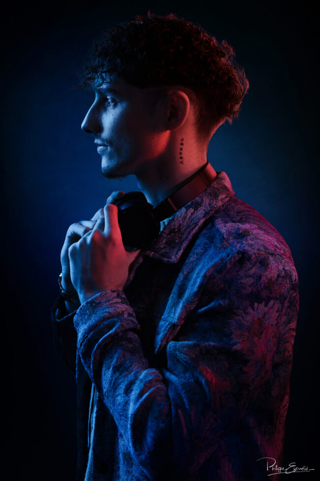 Portrait de profil d’un homme en veste texturée et casque autour du cou, éclairé par un subtil mélange de lumières rouges et bleues.