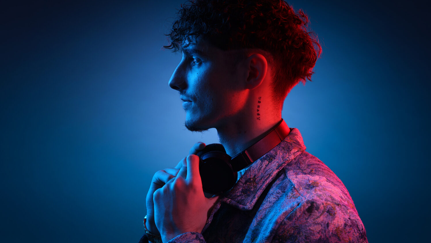 Portrait professionnel en profil d’un homme portant un casque audio autour du cou, capturé sous une lumière mixte rouge et bleue dans le studio avec signature en bas à droite philippe escudié