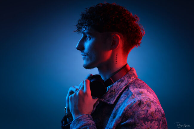 Portrait professionnel en profil d’un homme portant un casque audio autour du cou, capturé sous une lumière mixte rouge et bleue dans le studio avec signature en bas à droite philippe escudié