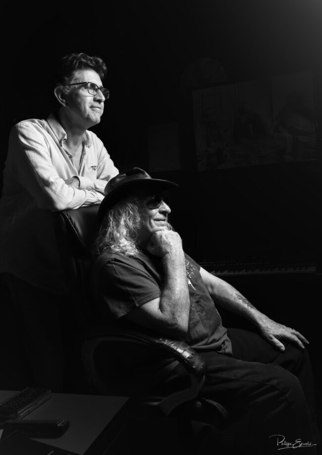 Portrait en noir et blanc de deux hommes complices dans un studio, l’un assis pensif avec un chapeau, l’autre debout derrière lui, ambiance cinématographique.