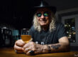 Portrait couleur d’un homme souriant avec chapeau et lunettes, assis à un comptoir avec un verre de bière, atmosphère chaleureuse et décontractée.