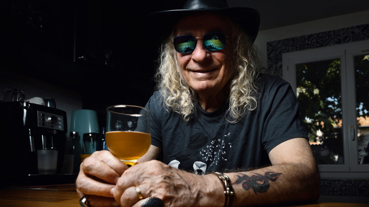 Portrait couleur d’un homme souriant avec chapeau et lunettes, assis à un comptoir avec un verre de bière, atmosphère chaleureuse et décontractée.