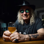 Portrait couleur d’un homme souriant avec chapeau et lunettes, assis à un comptoir avec un verre de bière, atmosphère chaleureuse et décontractée.