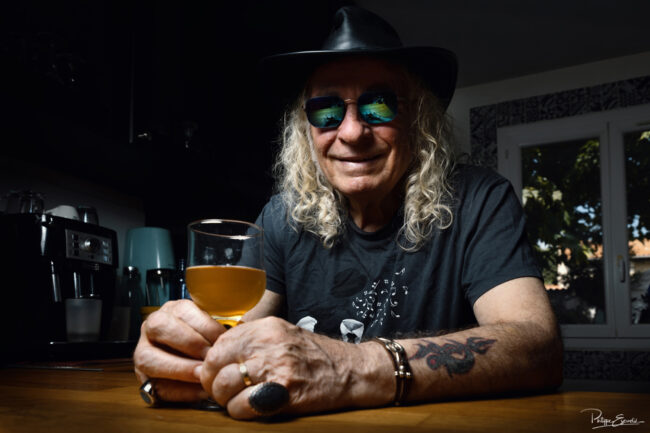 Portrait couleur d’un homme souriant avec chapeau et lunettes, assis à un comptoir avec un verre de bière, atmosphère chaleureuse et décontractée.