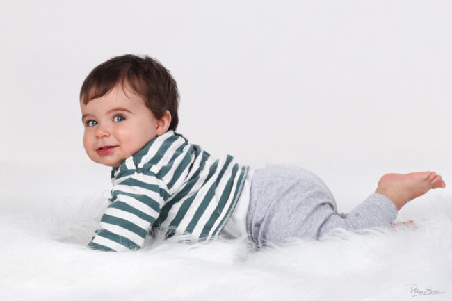 Bébé en tee shirt rayé allongé sur un tapis blanc qui regarde l’objectif en studio
