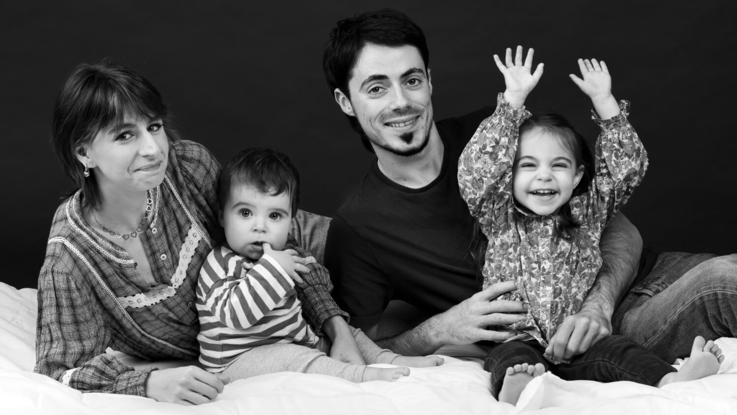 Portrait de famille souriante en noir et blanc posé sur un lit en studio, la famille est composée d'un homme , d'une femme, d'une petite fille et d'un bébé.