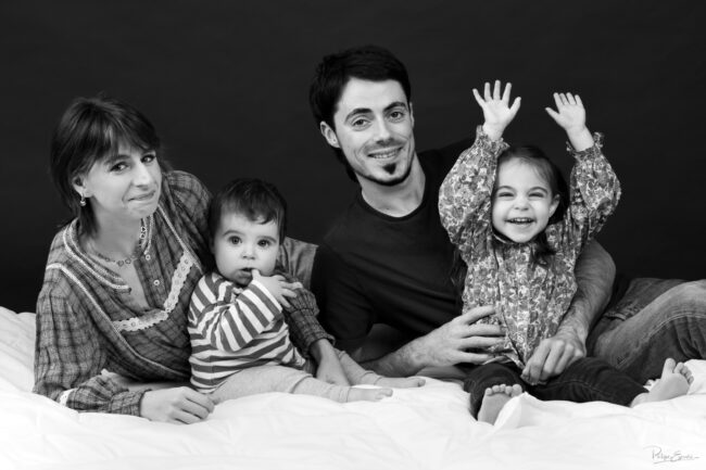 Portrait de famille souriante en noir et blanc posé sur un lit en studio, la famille est composée d'un homme , d'une femme, d'une petite fille et d'un bébé.