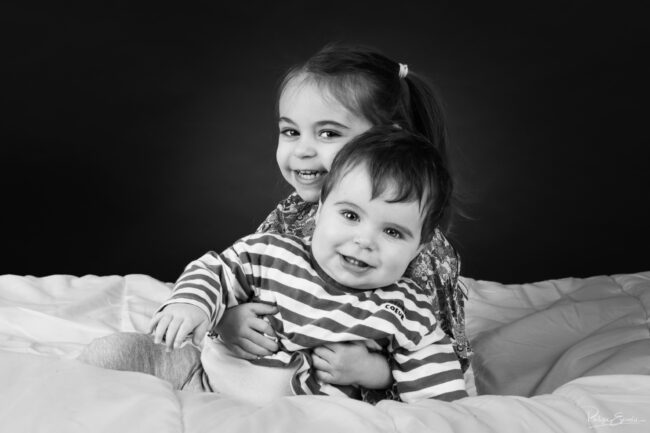 Frère et sœur enlacés et souriants sur fond noir en studio photo noir et blanc