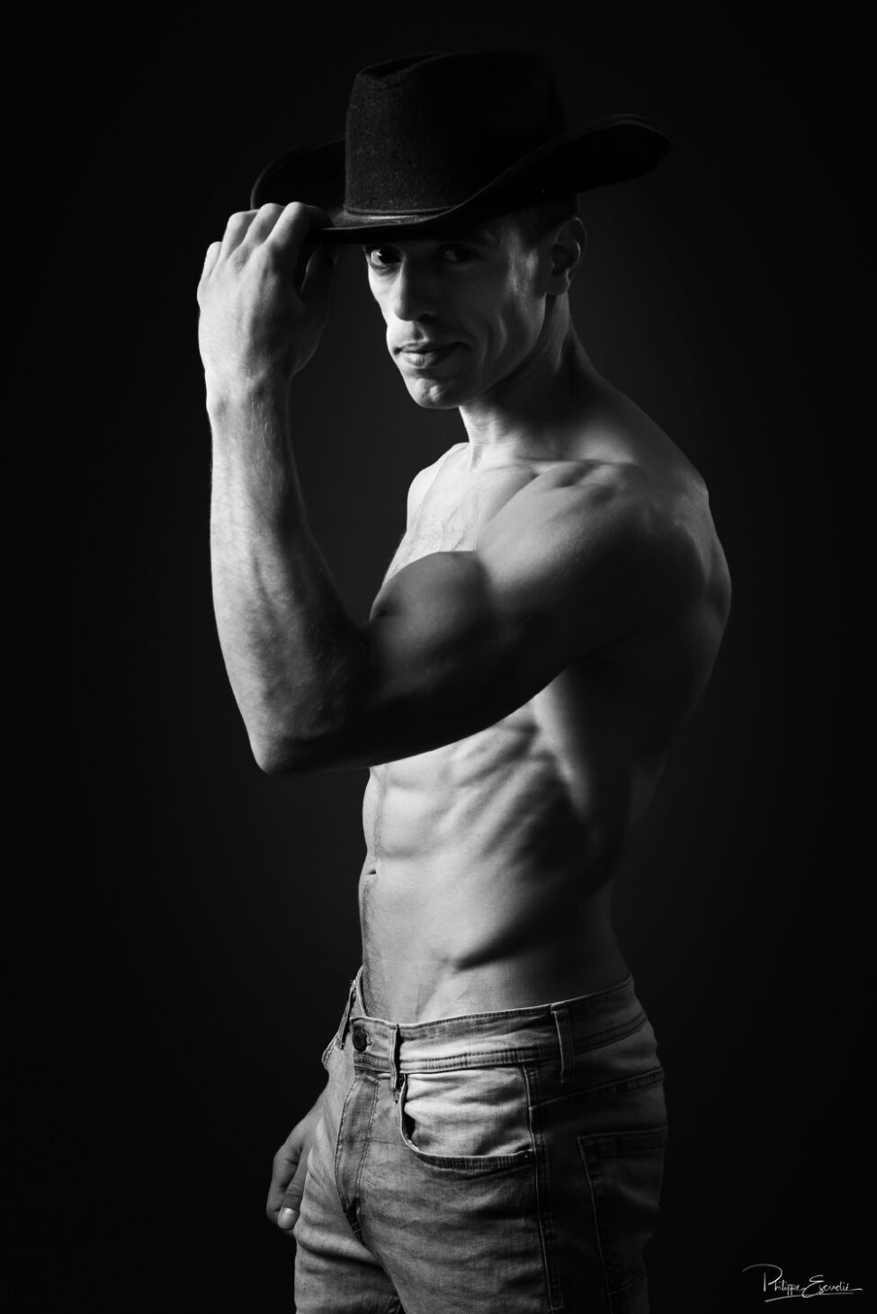 Portrait noir et blanc d’un homme torse nu portant un chapeau, éclairage contrasté en studio.