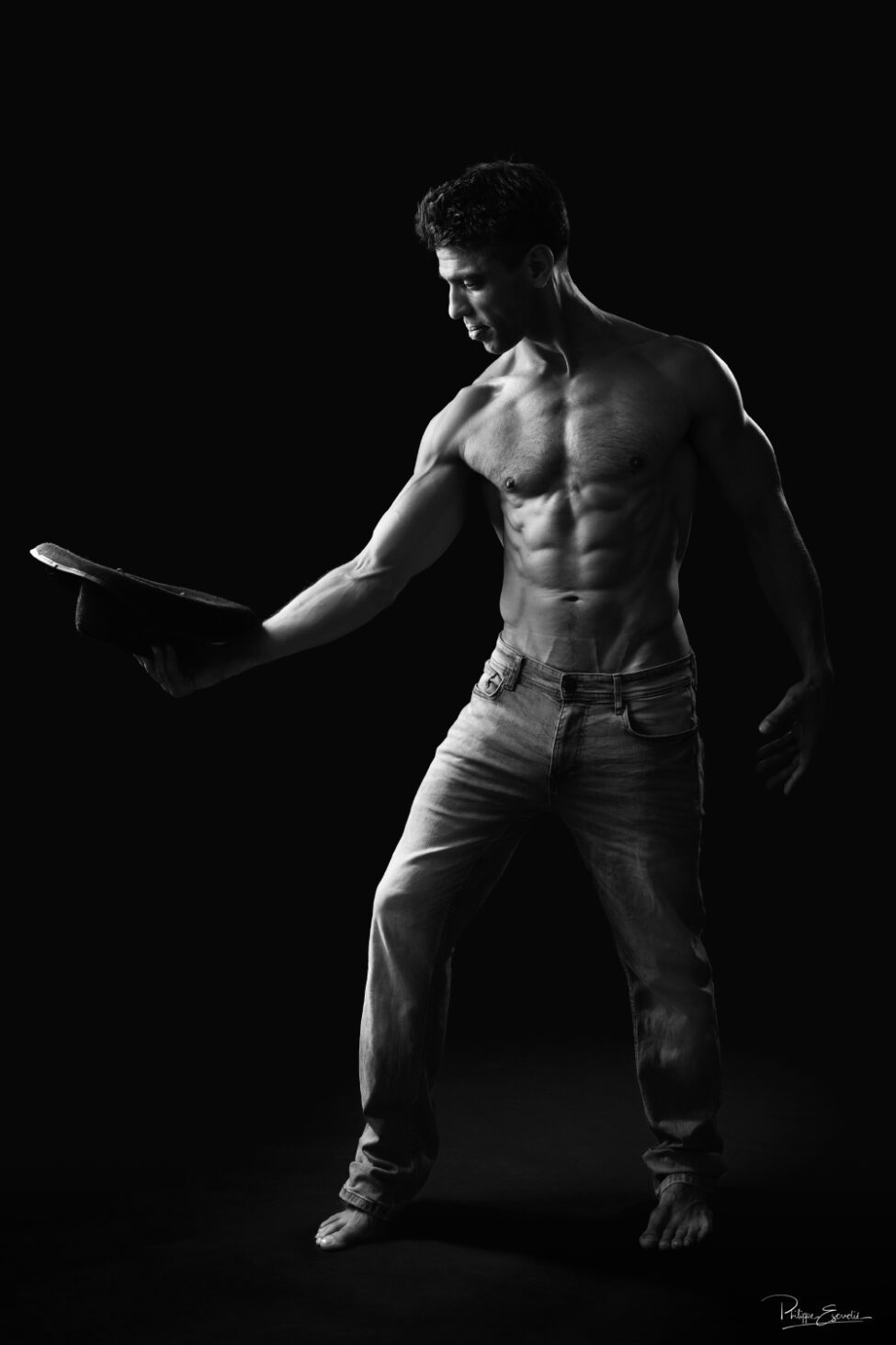 Portrait en noir et blanc mettant en valeur la silhouette athlétique d’un homme tenant un chapeau.