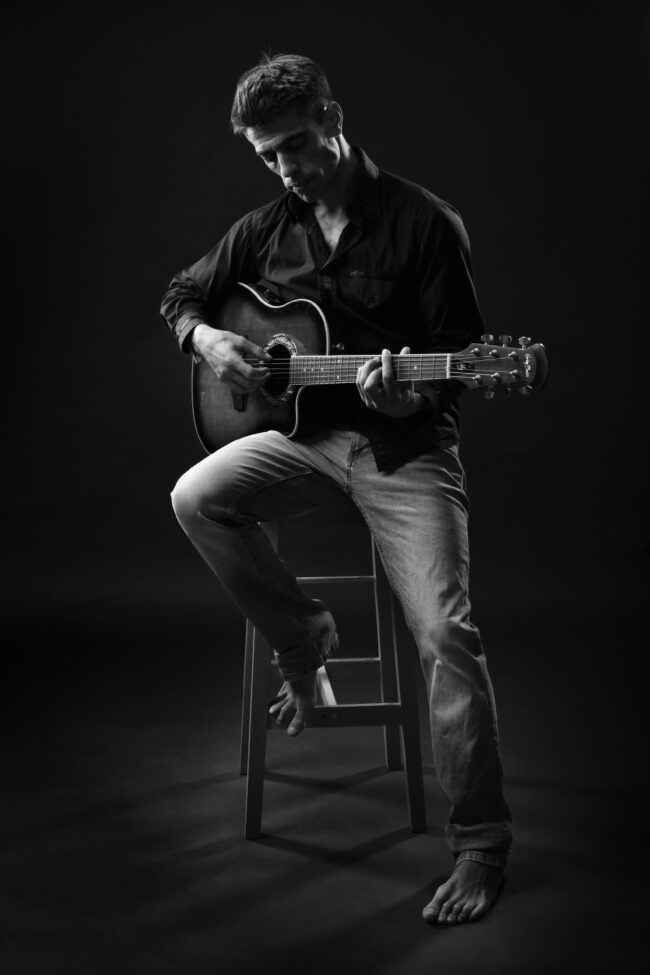Portrait d’un homme jouant de la guitare en studio, ambiance intimiste en noir et blanc.