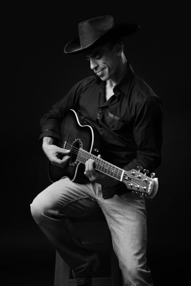 Un homme assis sur une chaise joue de la guitare avec un chapeau sur la tête, image en noir et blanc.