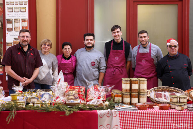 Portrait d’équipe derrière un stand, avec produits alimentaires exposés et façade de boutique en arrière-plan.