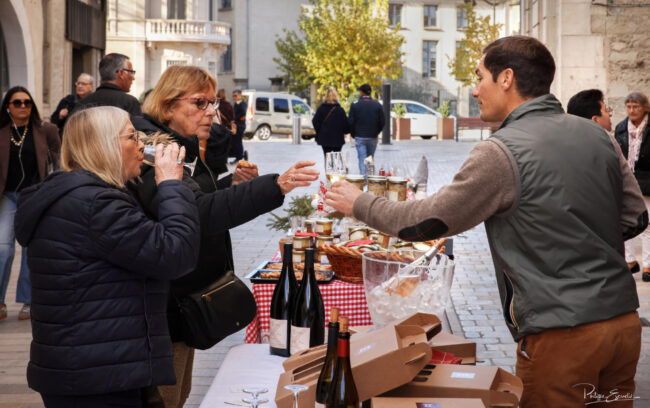 Scène de dégustation autour d’un stand en extérieur, avec échange de verres et produits posés sur la table.