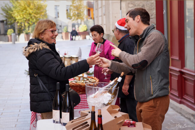 Scène de dégustation autour d’un stand alimentaire en extérieur, avec échange de verres et bouteilles sur la table.