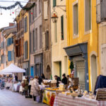 Stands commerciaux installés le long d’une rue piétonne, avec passants, tables de produits et décorations visibles en hauteur.