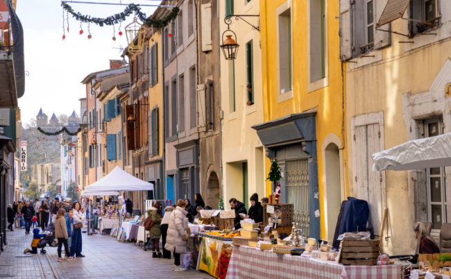 Stands commerciaux installés le long d’une rue piétonne, avec passants, tables de produits et décorations visibles en hauteur.