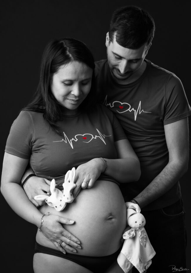 Un couple pose les mains sur le ventre arrondi tandis qu’un petit doudou évoque l’arrivée prochaine du bébé.
