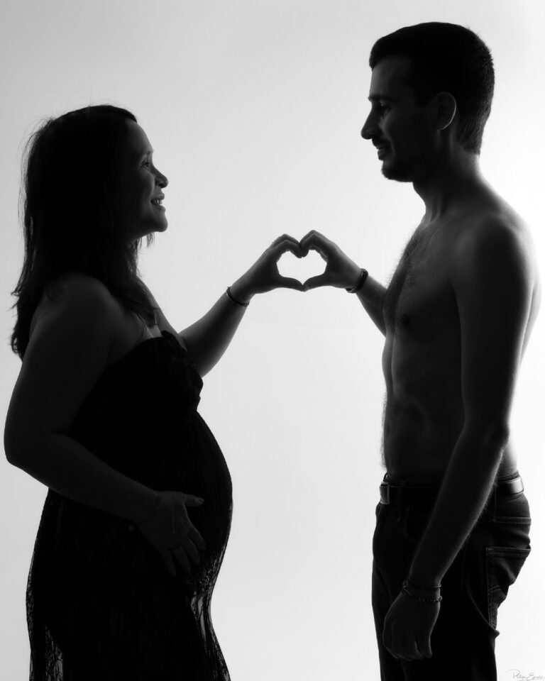 Shooting grossesse au studio photo de Carcassonne – Mai Lynh et Thomas 1 Silhouette d’un couple face à face formant un cœur avec leurs mains, la maman est enceinte et pose son autre main sur son ventre arrondi.