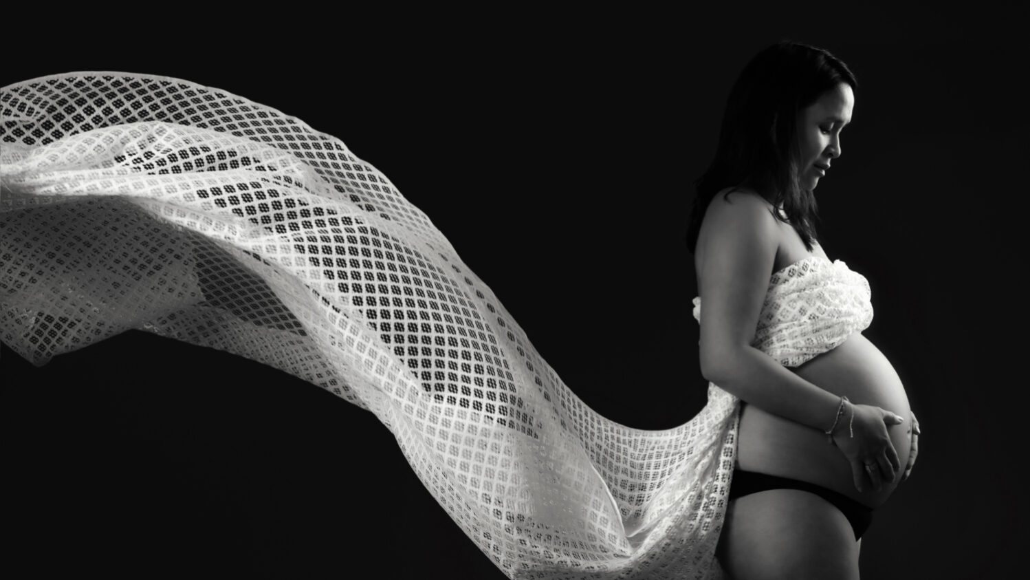 Portrait de grossesse en clair-obscur noir et blanc où un voile léger se déploie derrière la future maman, créant une image à la fois douce et graphique.
