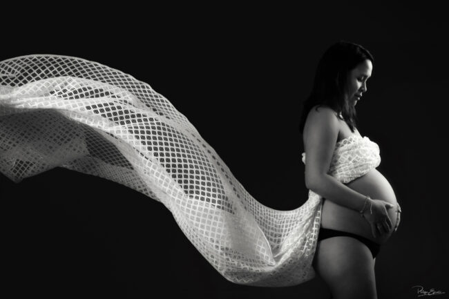 Portrait de grossesse en clair-obscur noir et blanc où un voile léger se déploie derrière la future maman, créant une image à la fois douce et graphique.