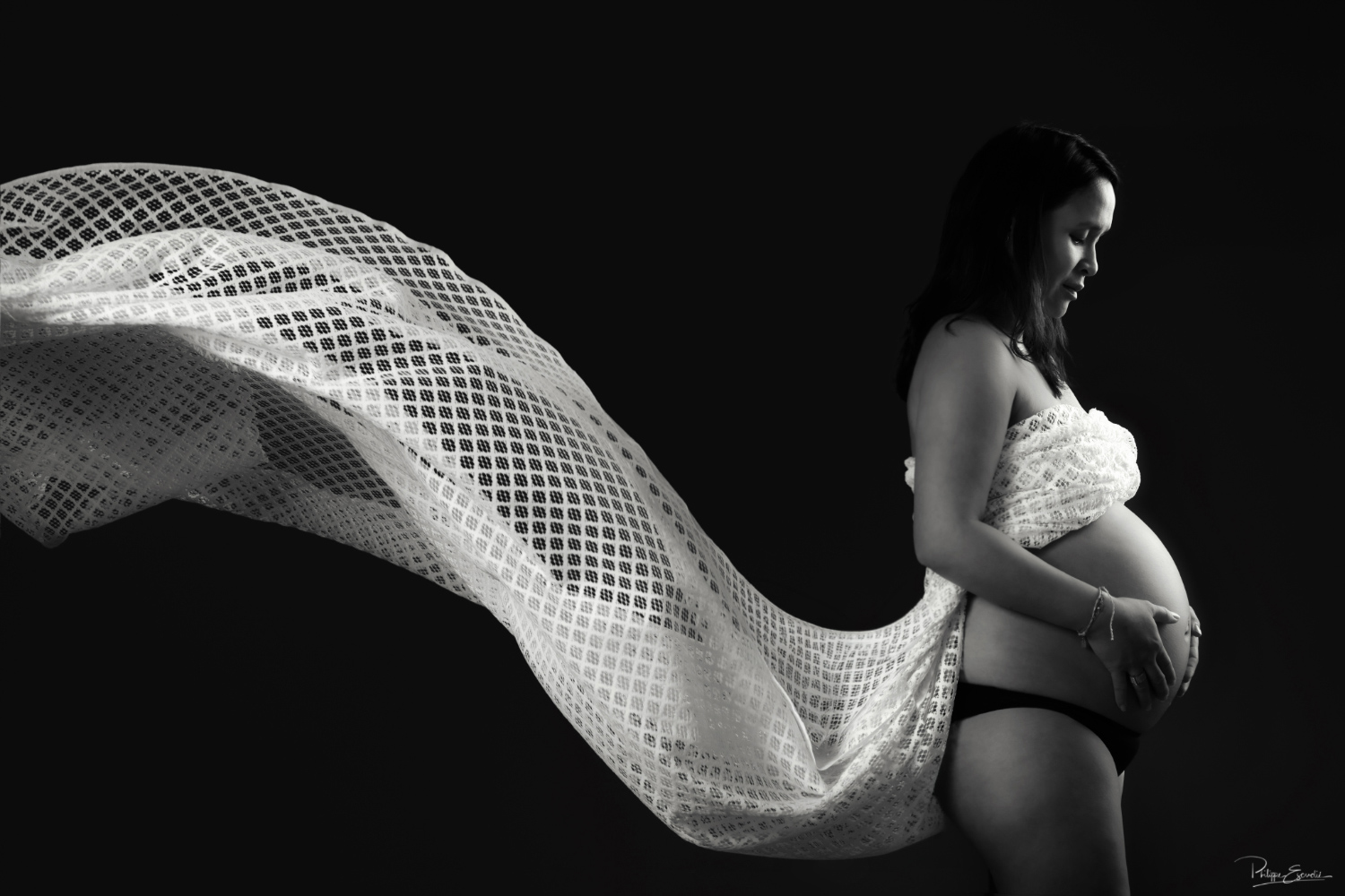 photo-grossesse-studio-voile-en-mouvement Portrait de grossesse en clair-obscur noir et blanc où un voile léger se déploie derrière la future maman, créant une image à la fois douce et graphique.