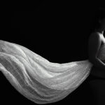 Portrait de grossesse en clair-obscur où un voile léger se déploie derrière la future maman, créant une image à la fois douce et graphique en noir et blanc.