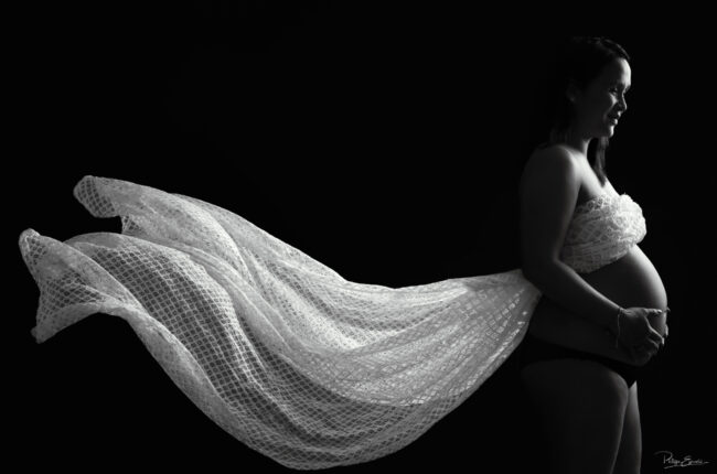 Portrait de grossesse en clair-obscur où un voile léger se déploie derrière la future maman, créant une image à la fois douce et graphique en noir et blanc.