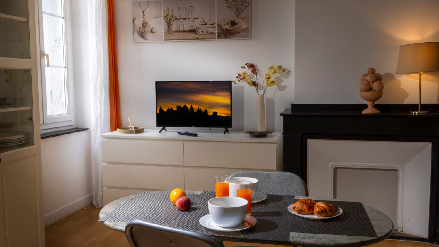 Ambiance chaleureuse et éclairage tamisé dans une pièce de vie, avec un petit déjeuner sur une table et un téléviseur en arrière plan sur lequel on voit la cité de Carcassonne au crépuscule.
