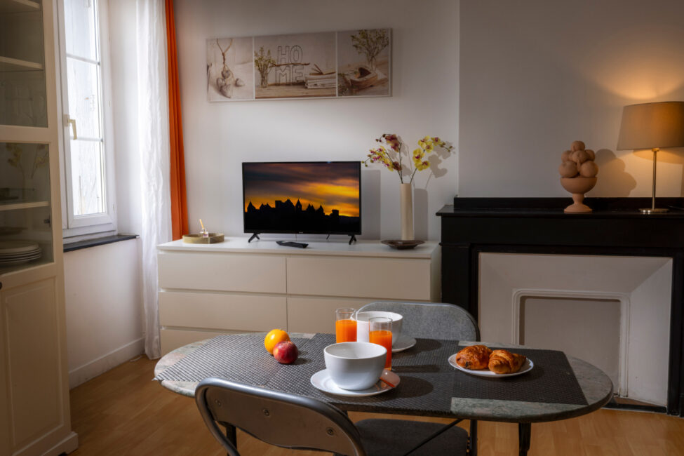 Ambiance chaleureuse et éclairage tamisé dans une pièce de vie, avec un petit déjeuner sur une table et un téléviseur en arrière plan sur lequel on voit la cité de Carcassonne au crépuscule.