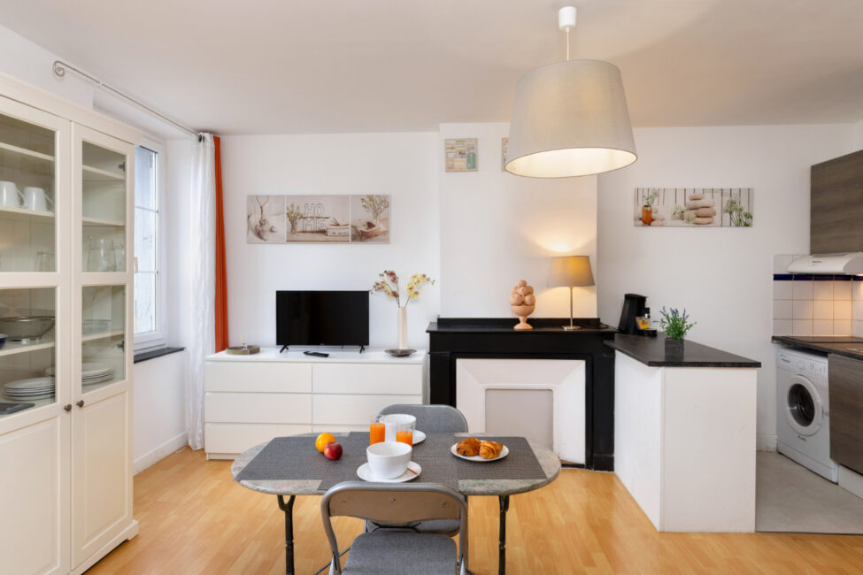 Salle à manger avec cheminée décorative, table avec petit déjeuner dressé, cuisine ouverte et décoration épurée dans un appartement rénové.
